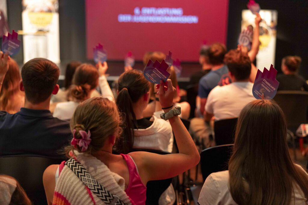 Eine Gruppe von Personen, die in einem Auditorium sitzt, hebt lila Karten hoch und blickt auf eine Bühne, auf der ein roter Bildschirm mit Text angezeigt wird, der zur Teilnahme an einer Abstimmung oder einer interaktiven Veranstaltung auffordert.