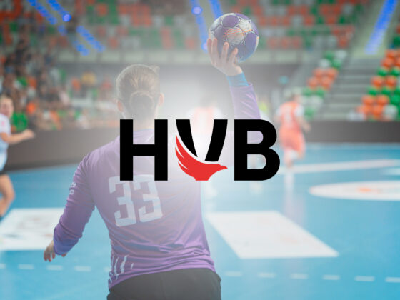 Ein Handballspieler im lila Trikot mit der Nummer 33 bereitet sich während eines Handballspiels in Brandenburg in einer Halle auf den Wurf vor. Im Hintergrund sind Zuschauer und Spieler zu sehen. Der Schriftzug HVB mit einem roten Flügellogo ist über das Bild gelegt.