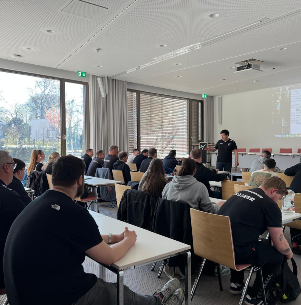Eine Gruppe von Personen sitzt an Tischen in einem hellen Konferenzraum und hört einem Redner zu, der vorne neben einer Leinwand mit einer projizierten Präsentation steht. Große Fenster lassen natürliches Licht herein.