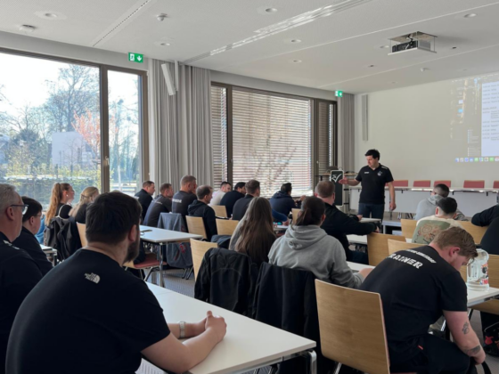 Eine Gruppe von Personen sitzt an Tischen in einem hellen Konferenzraum und hört einem Redner zu, der vorne neben einer Leinwand mit einer projizierten Präsentation steht. Große Fenster lassen natürliches Licht herein.