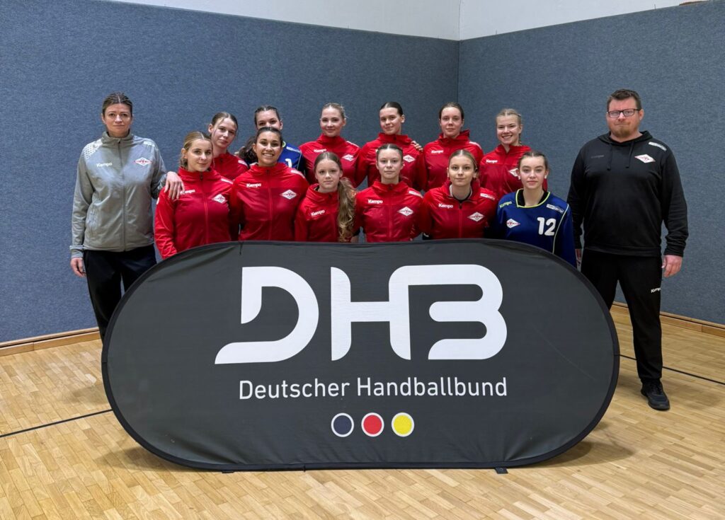 Eine Jugendhandballmannschaft in roten Trikots posiert in einer Halle hinter einem Banner des Deutschen Handballbundes mit zwei Trainern, die auf einem Holzboden vor einer grau-weißen Wand stehen.