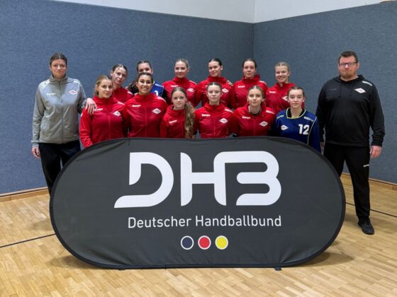 Eine Jugendhandballmannschaft in roten Trikots posiert in einer Halle hinter einem Banner des Deutschen Handballbundes mit zwei Trainern, die auf einem Holzboden vor einer grau-weißen Wand stehen.