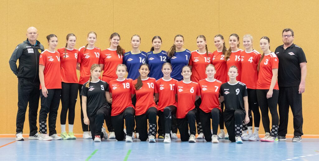Eine Mädchenhandballmannschaft stellt sich in einer Halle für ein Gruppenfoto mit zwei Trainern auf. Die meisten Spielerinnen tragen rote Trikots und schwarze Hosen, während zwei in der Mitte blaue Trikots tragen. Sie stehen alle auf dem Boden einer Sporthalle vor einer hellbraunen Wand.