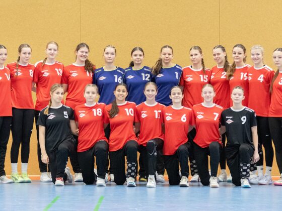 Eine Mädchenhandballmannschaft stellt sich in einer Halle für ein Gruppenfoto mit zwei Trainern auf. Die meisten Spielerinnen tragen rote Trikots und schwarze Hosen, während zwei in der Mitte blaue Trikots tragen. Sie stehen alle auf dem Boden einer Sporthalle vor einer hellbraunen Wand.
