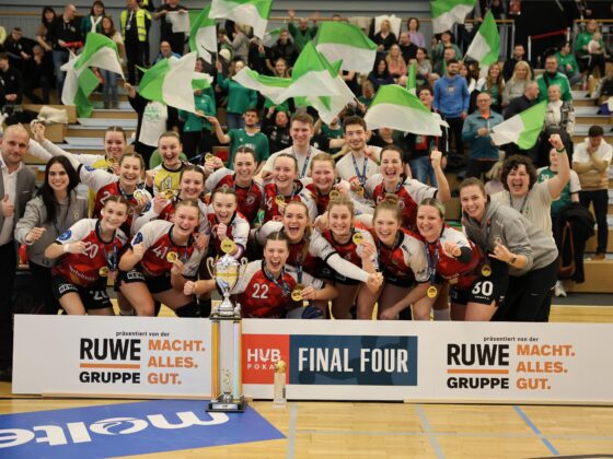 Eine Frauenhandballmannschaft in roten Trikots feiert mit einer Trophäe vor einem "FINAL FOUR"-Banner, lächelnd und jubelnd. Im Hintergrund stehen Fans mit grünen und weißen Fahnen auf Tribünen.