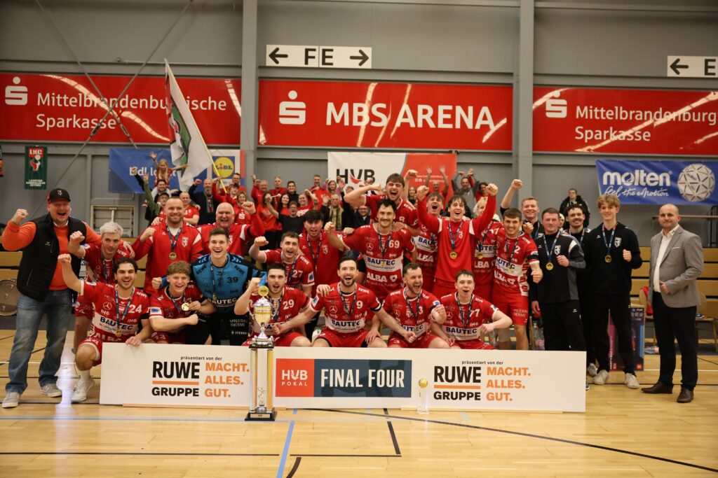 Ein jubelndes Männerhandballteam in roten Trikots feiert auf einem Sportplatz mit einem Pokal, hält Fäuste und Fahnen hoch, während Trainer und Betreuer daneben stehen. Im Hintergrund sind Meisterschaftsbanner und Sponsoren zu sehen.