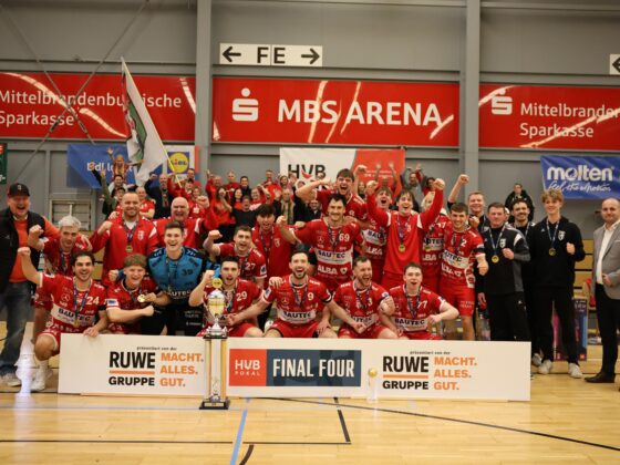 Ein jubelndes Männerhandballteam in roten Trikots feiert auf einem Sportplatz mit einem Pokal, hält Fäuste und Fahnen hoch, während Trainer und Betreuer daneben stehen. Im Hintergrund sind Meisterschaftsbanner und Sponsoren zu sehen.