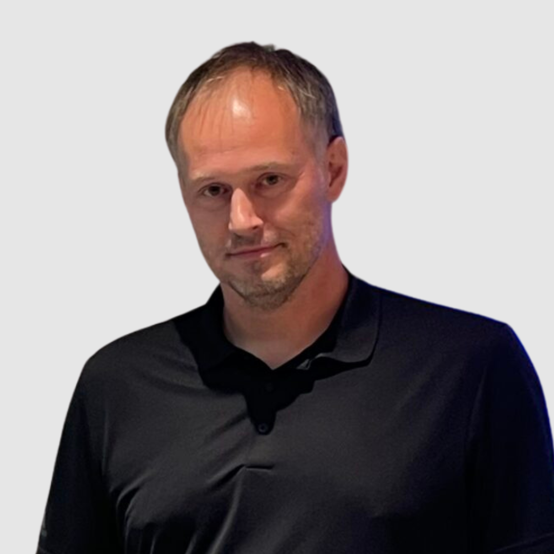 Ein Mann mit kurzen braunen Haaren, heller Gesichtsbehaarung und einem leichten Lächeln trägt ein schwarzes Hemd mit Kragen und steht vor einem schlichten hellgrauen Hintergrund.