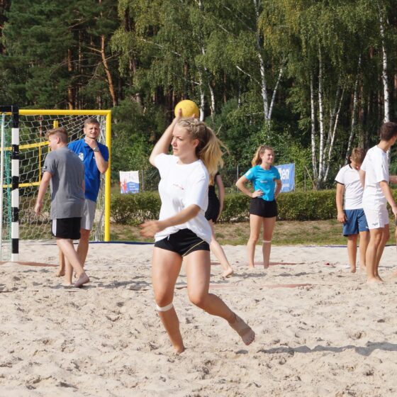 Eine Gruppe junger Leute spielt Beachhandball auf Sand in der Nähe eines Waldes. Ein Mädchen im Vordergrund bewegt sich und hält einen gelben Ball, während andere in der Nähe eines Torpfostens im Hintergrund stehen, reden oder zusehen.