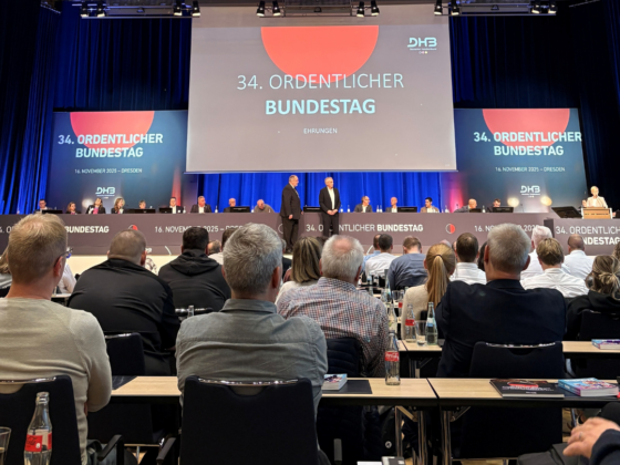 Ein großer Konferenzraum, gefüllt mit Menschen, die einer Bühne gegenüberstehen. Auf der Bühne befinden sich eine Reihe von Rednern und große Bildschirme, auf denen 34. Ordentlicher Bundestag. Auf den Tischen im Publikum stehen Flaschen und Notizbücher.