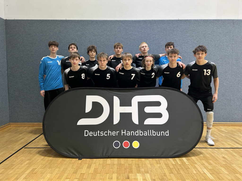 Eine Jungen-Handballmannschaft in schwarz-blauen Trikots posiert hinter einem Banner des Deutschen Handballbundes in einer Sporthalle mit Holzboden und grauer Wand.