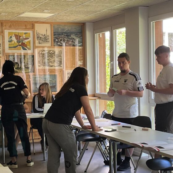 Mehrere junge Erwachsene arbeiten zusammen in einem hellen Klassenzimmer, stehen oder sitzen mit Papier und Stiften um Tische herum. Im Hintergrund sind Kunstwerke und Fenster mit Grünpflanzen zu sehen.