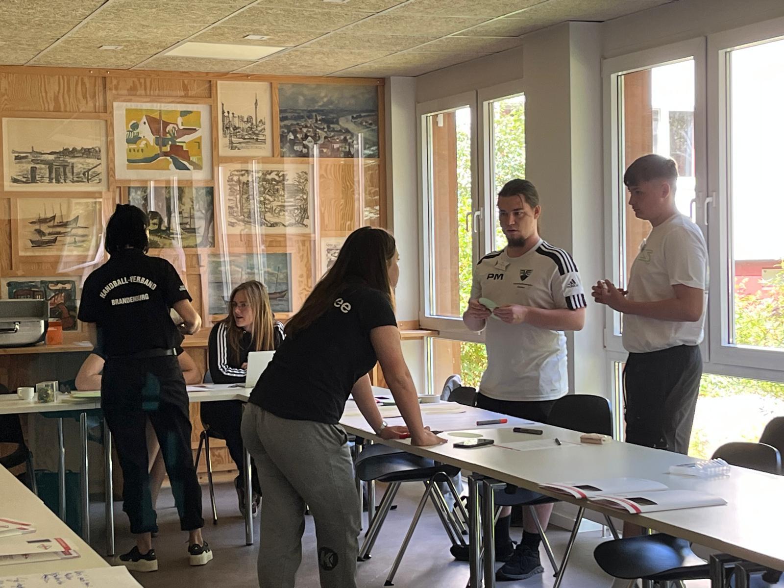 Mehrere junge Erwachsene arbeiten zusammen in einem hellen Klassenzimmer, stehen oder sitzen mit Papier und Stiften um Tische herum. Im Hintergrund sind Kunstwerke und Fenster mit Grünpflanzen zu sehen.
