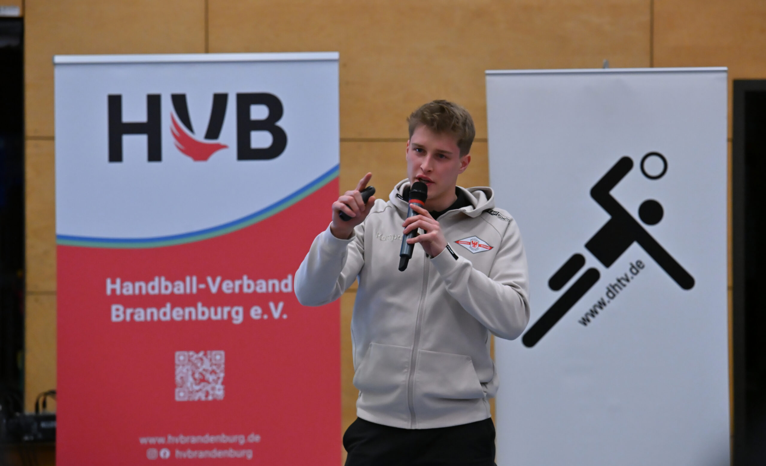Ein junger Mann in einem hellgrauen Kapuzenpulli spricht in ein Mikrofon und gestikuliert dabei mit der Hand. Hinter ihm sind Banner des Handball-Verbandes Brandenburg e.V. und ein stilisiertes Handballer-Logo zu sehen.