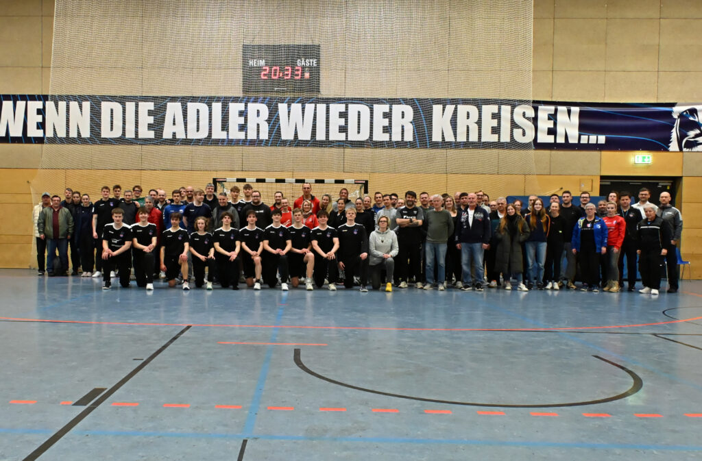 Eine große Gruppe von Menschen, darunter zwei Mannschaften in Sportuniformen, posieren gemeinsam in einer Turnhalle vor einem Transparent mit der Aufschrift "WENN DIE ADLER WIEDER KREISEN...". Der Boden ist mit Markierungen für ein Sportfeld versehen.