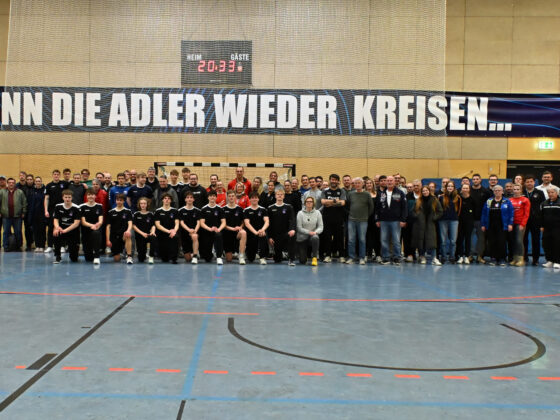 Eine große Gruppe von Menschen, darunter zwei Mannschaften in Sportuniformen, posieren gemeinsam in einer Turnhalle vor einem Transparent mit der Aufschrift "WENN DIE ADLER WIEDER KREISEN...". Der Boden ist mit Markierungen für ein Sportfeld versehen.