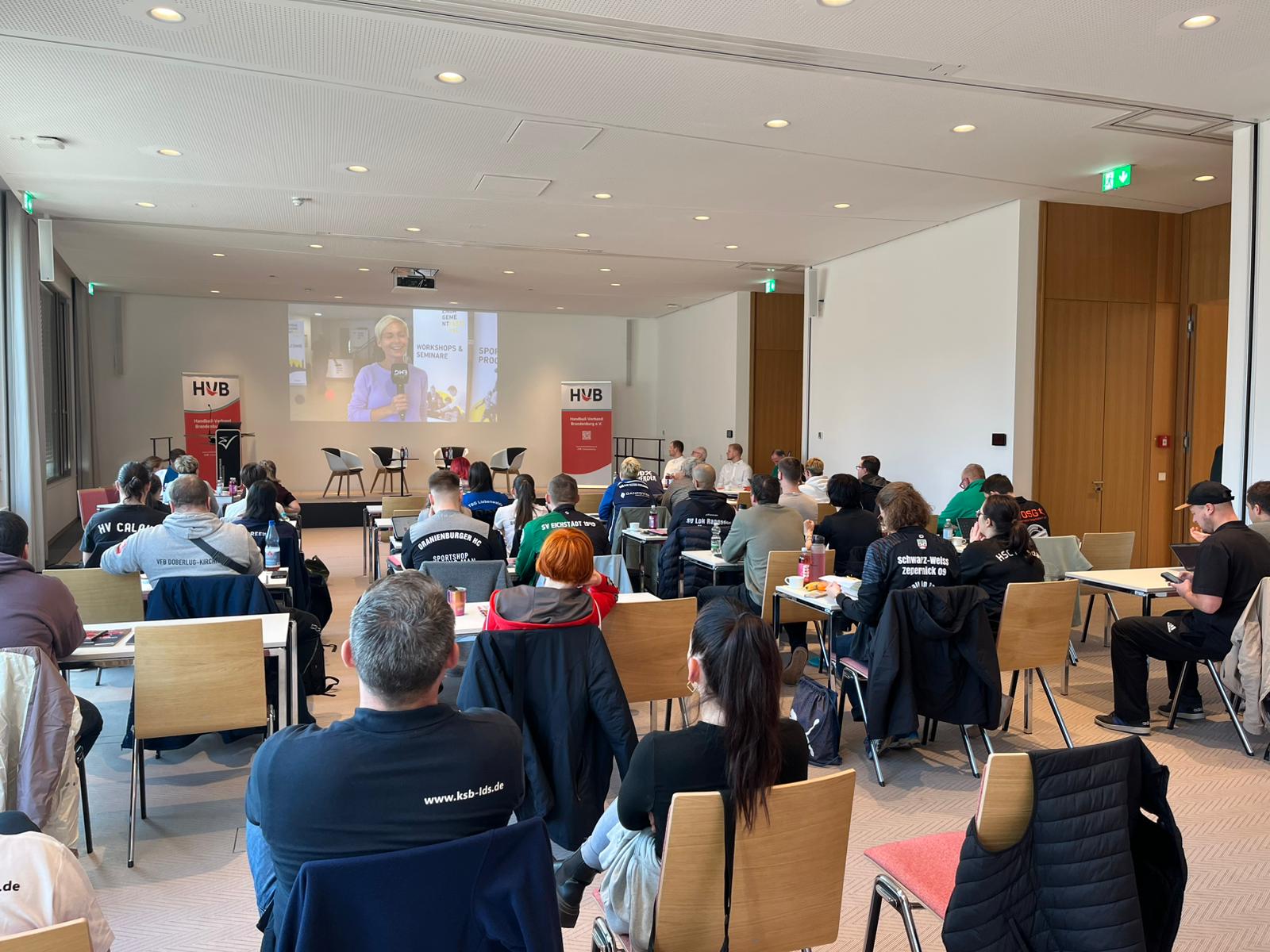 In einem Konferenzraum sitzen Menschen vor einem Bildschirm, auf dem eine Frau eine Präsentation hält. Auf zwei Bannern steht HVB. Der Raum hat Holzböden, große Fenster und eine Mischung aus Männern und Frauen.