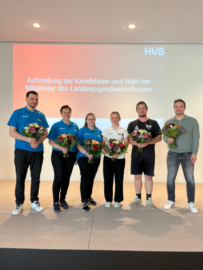 Sechs Personen stehen auf einer Bühne, vier davon mit Blumensträußen in der Hand, vor einem Bildschirm, auf dem ein deutscher Text über die Wahl der Mitglieder des Landesjugendausschusses angezeigt wird. Alle sind leger gekleidet und lächeln.
