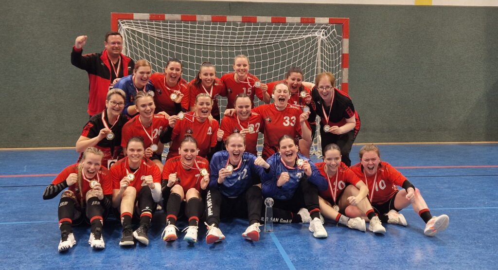 Eine Frauenhandballmannschaft in roten Trikots sitzt und kniet gemeinsam vor einem Tor, lächelt und jubelt, während sie ihre Medaillen für ein Gruppenfoto in einer Halle hochhält.