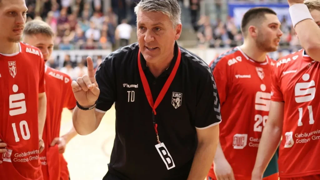 Ein Trainer in einem schwarzen Hemd mit einer Pfeife und einem Schlüsselband spricht während eines Spiels zu seiner Handballmannschaft in roten Trikots und hält einen Finger hoch, während die Spieler aufmerksam zuhören. Im Hintergrund sieht eine Menschenmenge zu.