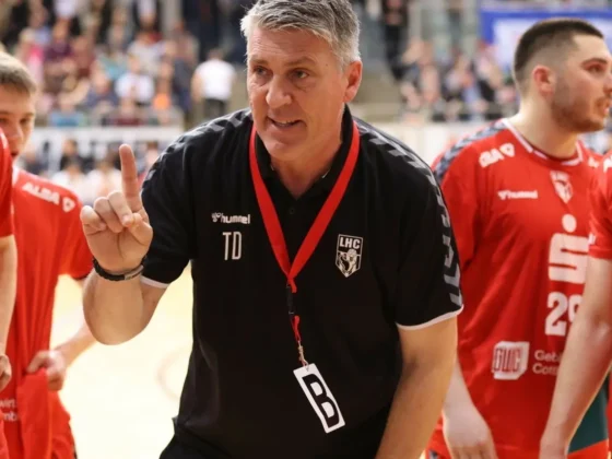 Ein Trainer in einem schwarzen Hemd mit einer Pfeife und einem Schlüsselband spricht während eines Spiels zu seiner Handballmannschaft in roten Trikots und hält einen Finger hoch, während die Spieler aufmerksam zuhören. Im Hintergrund sieht eine Menschenmenge zu.