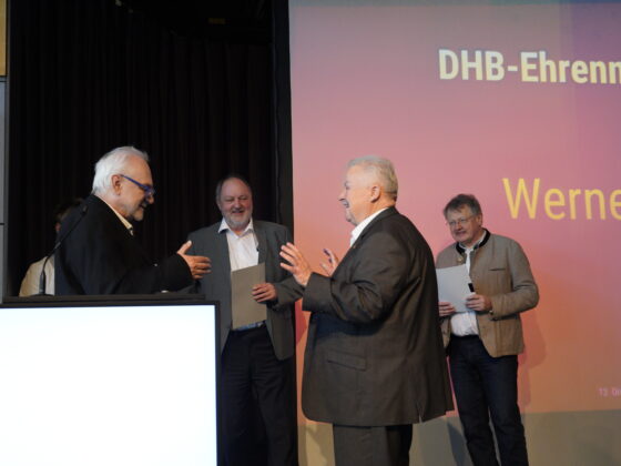 Vier Männer in Anzügen stehen während einer Preisverleihung für Handball in Brandenburg auf der Bühne. Ein Mann überreicht einem anderen vor einer Leinwand, auf der DHB-Ehrennad und Werner zu sehen sind, eine Urkunde, während zwei andere in der Nähe Urkunden halten.