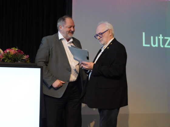 Zwei Männer in Anzügen stehen auf der Bühne, einer überreicht dem anderen ein Dokument, beide lächeln - vielleicht feiern sie einen Erfolg im Handball. Eine Frau mit Blumen steht an der Seite neben einem Podium, während "Lutz" teilweise auf dem Bildschirm hinter ihnen zu sehen ist.