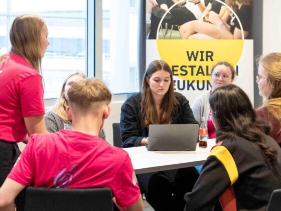 Eine Gruppe junger Erwachsener sitzt um einen Tisch und diskutiert über Handball. Eine Frau steht auf und wendet sich an die Gruppe. Hinter ihnen hängt ein Transparent mit Fotos und dem deutschen Text WIR GESTALTEN ZUKUNFT.