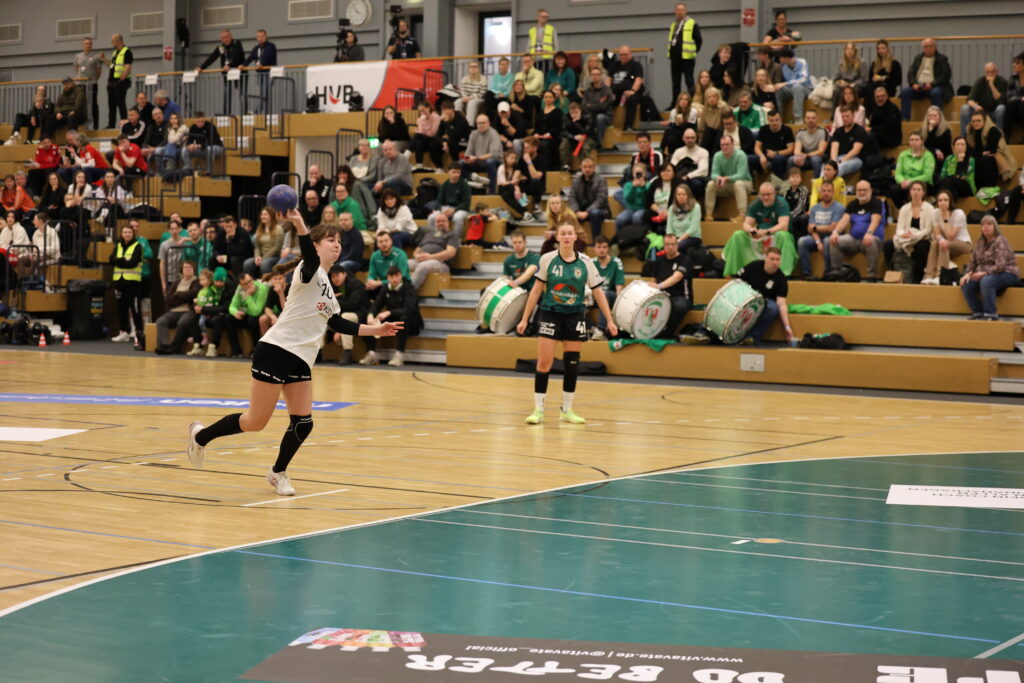 Eine Handballspielerin in einem weiß-schwarzen Trikot ist im Begriff, während eines Spiels einen Ball zu werfen. Eine andere Spielerin in Grün steht in der Nähe des Tores. Die Zuschauer auf der Tribüne im Hintergrund verfolgen das Geschehen.