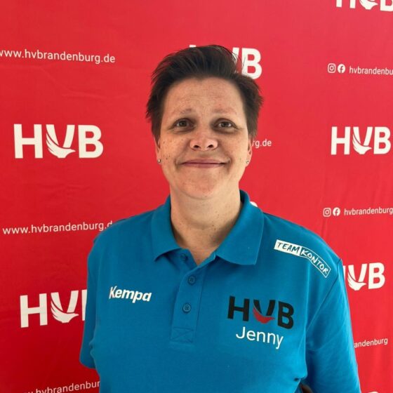 Eine Person mit kurzen dunklen Haaren, die ein blaues Poloshirt mit den Aufschriften Kempa, Team Kontor, Handball Brandenburg und Jenny trägt, steht lächelnd vor einem roten Hintergrund, auf dem wiederholt das HVB-Logo und die Website aufgedruckt sind.