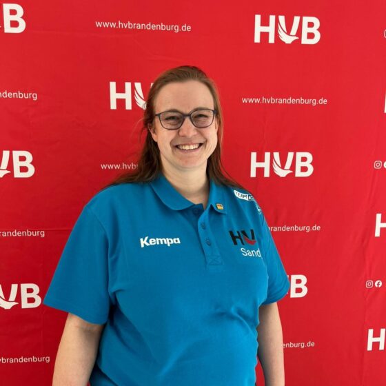 Eine lächelnde Person mit langen Haaren und Brille trägt ein blaues Poloshirt mit Logos und steht vor einem roten Handball-Brandenburg-Hintergrund, auf dem wiederholt HVB und hvbrandenburg.de aufgedruckt sind.
