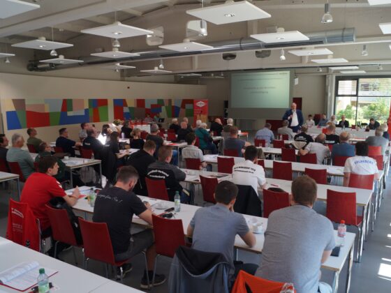 Eine Gruppe von Personen sitzt an Tischen in einem hellen, modernen Konferenzraum und hört einem Redner zu, der vorne neben einem Projektor steht. Die meisten Teilnehmer blicken nach vorne, einige machen sich Notizen.