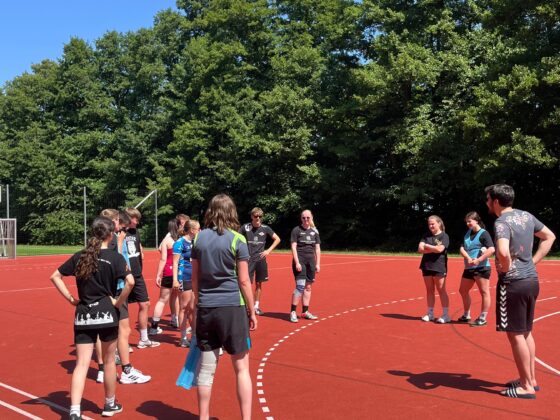 Eine Gruppe von Menschen steht in einem Halbkreis auf einem roten Sportplatz im Freien und hört einem Handballtrainer zu. Sie sind in Sportkleidung gekleidet, im Hintergrund sind Bäume und grüne Blätter unter einem klaren blauen Himmel zu sehen.