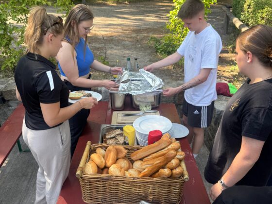 Vier Personen servieren das Essen im Freien an einem Picknicktisch, der mit Brötchen, Besteck, Tellern und einer abgedeckten Schale gedeckt ist. Nach einem freundschaftlichen Handballspiel entspannen sie sich gemeinsam inmitten von Bäumen und Grünpflanzen im Hintergrund.