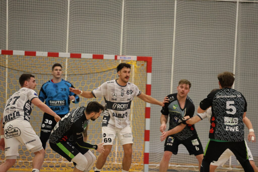 Mehrere männliche Handballspieler in Aktion in der Nähe des Tores, mit einem Torwart in Blau, der vor dem Netz steht. Die Spieler in weißen und schwarzen Trikots verteidigen und greifen während dieses intensiven Handballspiels aktiv an.