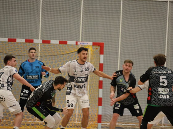 Mehrere männliche Handballspieler in Aktion in der Nähe des Tores, mit einem Torwart in Blau, der vor dem Netz steht. Die Spieler in weißen und schwarzen Trikots verteidigen und greifen während dieses intensiven Handballspiels aktiv an.