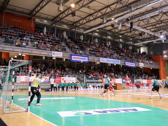 Ein spannendes Handballspiel findet in einer großen Halle mit Zuschauern auf der Tribüne statt. Die Spieler in grün-weißen Trikots konzentrieren sich auf das Geschehen in Tornähe, während Banner und Anzeigetafeln das spannende Handballspiel hervorheben.