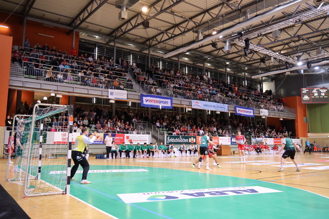 Ein spannendes Handballspiel findet in einer großen Halle mit Zuschauern auf der Tribüne statt. Die Spieler in grün-weißen Trikots konzentrieren sich auf das Geschehen in Tornähe, während Banner und Anzeigetafeln das spannende Handballspiel hervorheben.