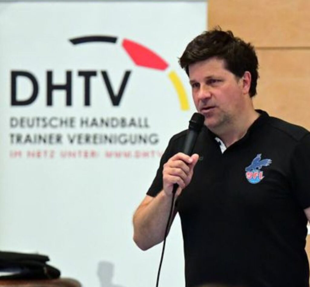 Ein Mann in einem schwarzen Hemd hält ein Mikrofon in der Hand und spricht über Handball vor einem Banner mit der Aufschrift DHTV Deutsche Handball Trainer Vereinigung. Die Umgebung in der Halle lässt vermuten, dass er eine Präsentation oder einen Vortrag hält.