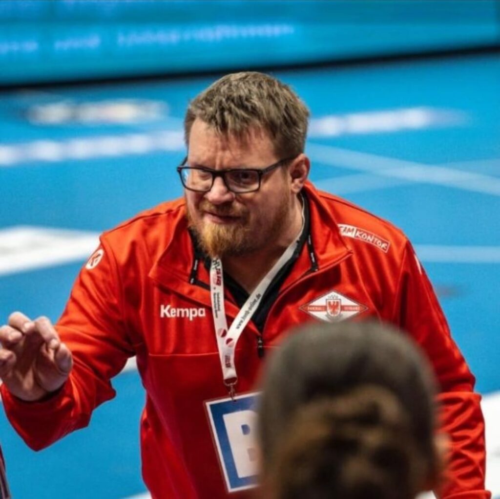 Ein Mann mit Brille und Bart, der eine rote Sportjacke und ein Schlüsselband trägt, gestikuliert mit seiner Hand. Er scheint zu trainieren oder Anweisungen zu geben, im Hintergrund ist ein unscharfes blaues Sportfeld zu sehen.