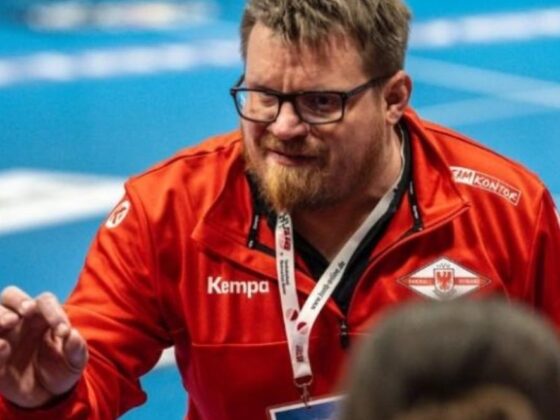 Ein Mann mit Brille und Bart, der eine rote Sportjacke und ein Schlüsselband trägt, gestikuliert mit seiner Hand. Er scheint zu trainieren oder Anweisungen zu geben, im Hintergrund ist ein unscharfes blaues Sportfeld zu sehen.