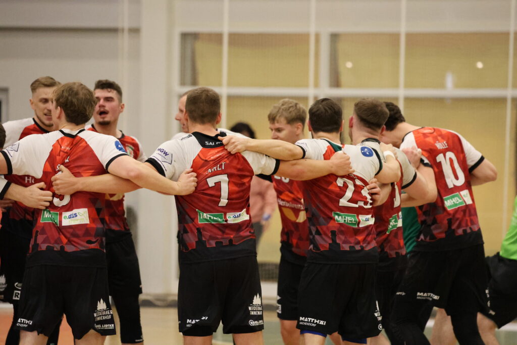Eine Gruppe von männlichen Sportlern in rot-schwarzen Trikots steht im Kreis und legt die Arme umeinander, um sich in der Halle gegenseitig zu feiern oder zu motivieren - ganz im Sinne des Nachwuchsleistungssports.