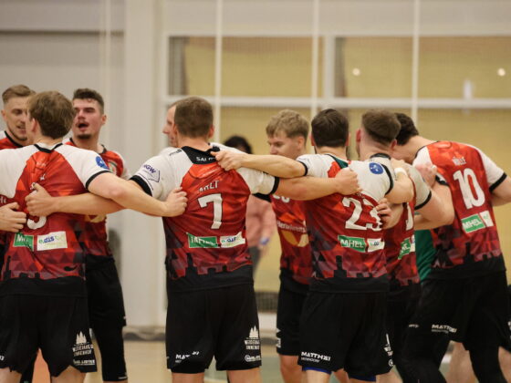 Eine Gruppe von männlichen Sportlern in rot-schwarzen Trikots steht im Kreis und legt die Arme umeinander, um sich in der Halle gegenseitig zu feiern oder zu motivieren - ganz im Sinne des Nachwuchsleistungssports.