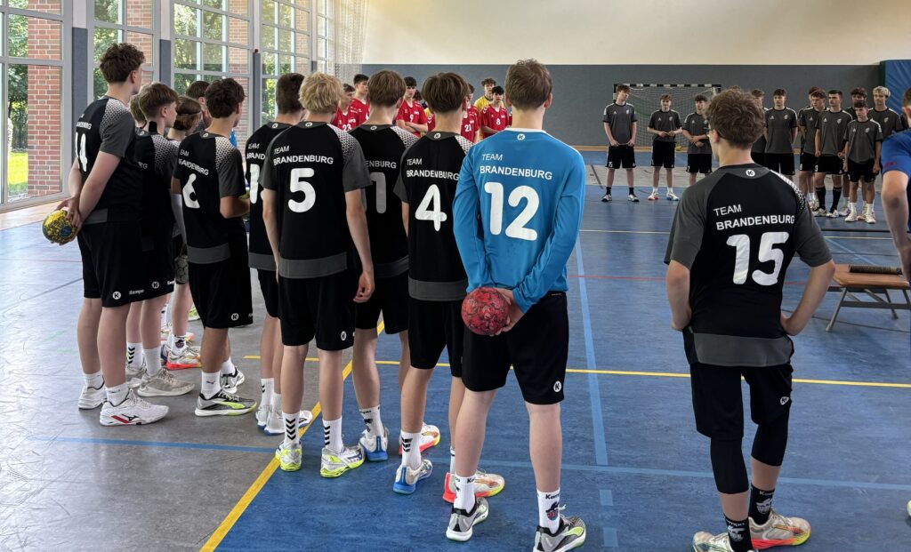 Eine Gruppe Jugendlicher in schwarzen Sportuniformen mit der Aufschrift "Team Brandenburg" steht in einer Turnhalle und bereitet sich auf ein Handballspiel gegen ein anderes Team in roten Uniformen vor.