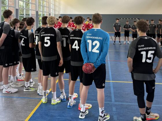 Eine Gruppe Jugendlicher in schwarzen Sportuniformen mit der Aufschrift "Team Brandenburg" steht in einer Turnhalle und bereitet sich auf ein Handballspiel gegen ein anderes Team in roten Uniformen vor.