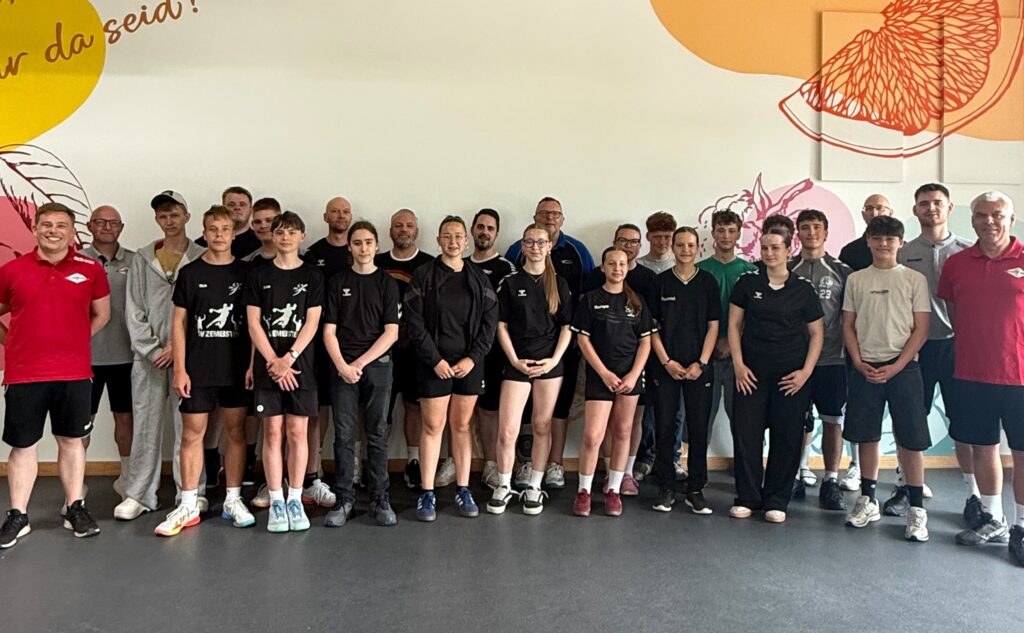 Eine Gruppe von etwa 25 Personen, darunter Erwachsene und Jugendliche, die Nachwuchsleistungssport betreiben, steht in Sportkleidung in einer Halle und lächelt in die Kamera. Im Hintergrund sind bunte Wandbilder mit Obstillustrationen und motivierenden Texten zu sehen.