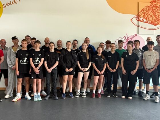Eine Gruppe von etwa 25 Personen, darunter Erwachsene und Jugendliche, die Nachwuchsleistungssport betreiben, steht in Sportkleidung in einer Halle und lächelt in die Kamera. Im Hintergrund sind bunte Wandbilder mit Obstillustrationen und motivierenden Texten zu sehen.