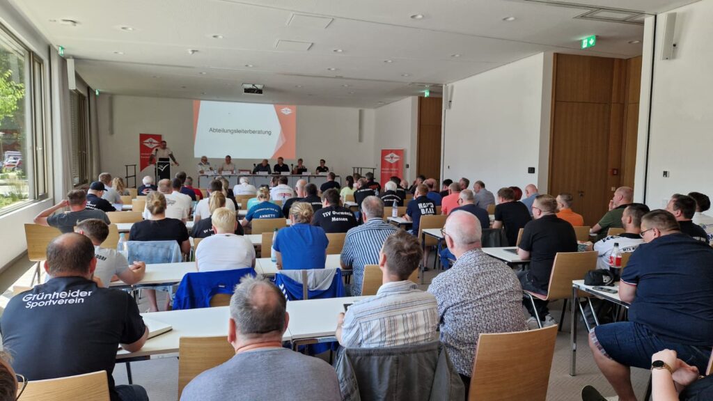 Eine große Gruppe von Menschen sitzt in Reihen vor einem Podium im vorderen Teil eines Konferenzraums und sieht sich einen auf eine Leinwand projizierten Vortrag über Nachwuchsleistungssport an. Einige Teilnehmer tragen Shirts mit den Namen ihrer Organisationen.