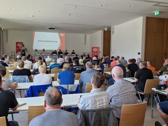 Eine große Gruppe von Menschen sitzt in Reihen vor einem Podium im vorderen Teil eines Konferenzraums und sieht sich einen auf eine Leinwand projizierten Vortrag über Nachwuchsleistungssport an. Einige Teilnehmer tragen Shirts mit den Namen ihrer Organisationen.