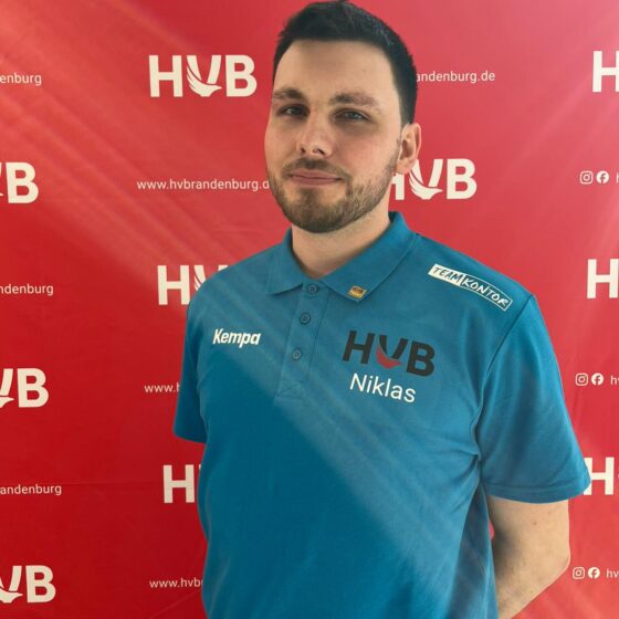 Ein Mann mit kurzen dunklen Haaren und einem gepflegten Bart steht vor einem roten Hintergrund, auf dem „HVB“ und „hvbrandenburg.de“ aufgedruckt sind. Er trägt ein blaues Poloshirt mit der Aufschrift „HVB Niklas“ und blickt leicht zur Seite.
